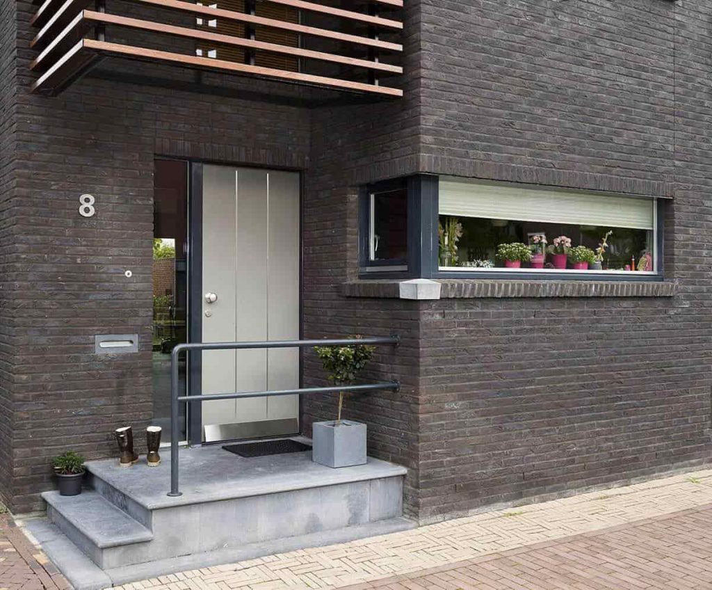 A contemporary black timber front door with a metallic handle, set in a dark brick facade with modern architectural elements. The design complements the urban exterior and showcases a minimalist, modern home entrance.