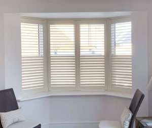 A charming bay window with white plantation shutters set in a light-filled room. The window seat area is accompanied by two chairs, offering a comfortable place to relax and enjoy the natural light that streams through the adjustable shutter slats.