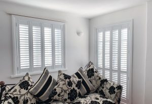 A cosy nook with a plush sofa adorned with black and white floral-patterned cushions. The window behind the sofa is fitted with white plantation shutters, creating a contrast with the soft, neutral tones of the room's walls and the sofa's fabric.