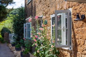 A picturesque stone cottage with Residence 7 timber effect windows, beautifully framed by blooming flowers and rustic stone walls, under a clear blue sky.