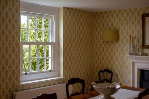 uPVC sliding sash window in a dining room with yellow patterned wallpaper, overlooking green trees outside.