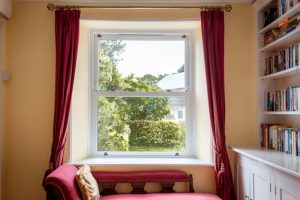  uPVC sliding sash window framed by red curtains in a cosy reading nook, overlooking a garden.