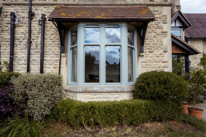 A bay window with uPVC sash frames set into a stone-walled house surrounded by shrubs.