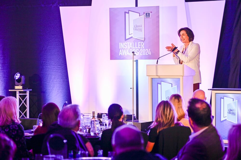 A presenter speaks at a podium during the Glass and Glazing Installer Awards 2024, addressing an engaged audience in a professionally lit venue with branded displays.